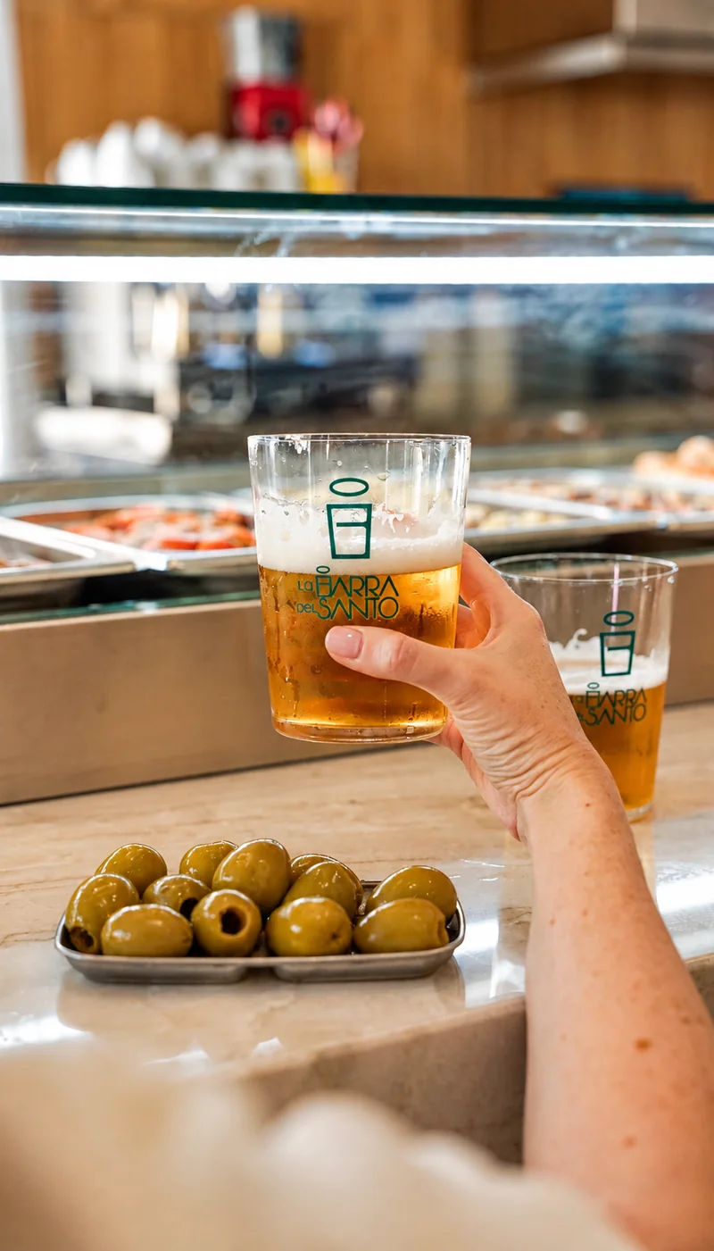 Una mano sostiene un vaso de cerveza sobre la barra, junto a un plato de aceitunas.