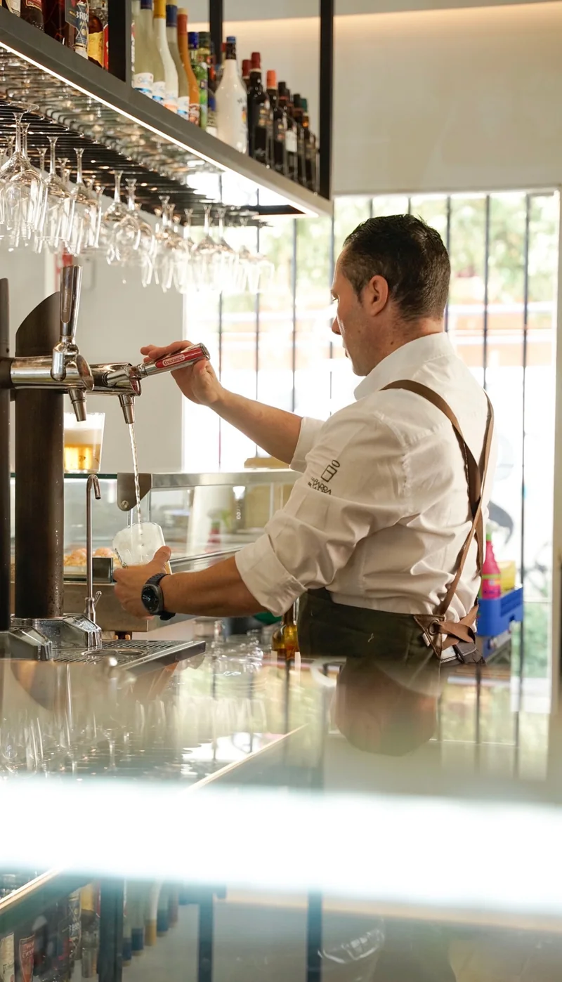 Persona sirviendo cerveza de grifo detrás de la barra, bajo un estante con copas.
